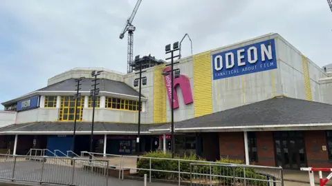 The picture shows the ODEON in Bracknell, the blue and white ODEON logo is visible on the top right of the building. It is a grey building with yellow wood panelling.  