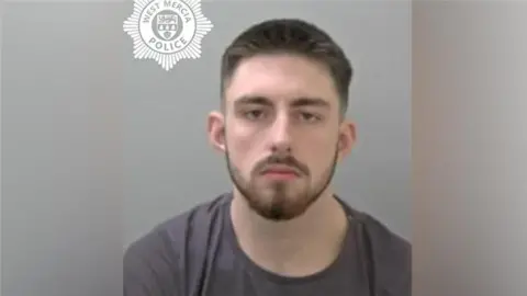 A mugshot of a man with brown hair and brown facial hair. He is wearing a dark grey t-shirt