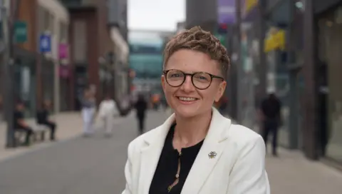 Bev Craig has short red hair, and is wearing glasses and a white jacket over a black blouse