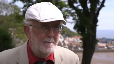 An image of Councillor Randerson in a cream cap, glasses and a cream suit in front of Scarborouugh South Bay