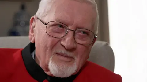 An elderly man with silver hair and beard, wearing glasses, smiles wistfully