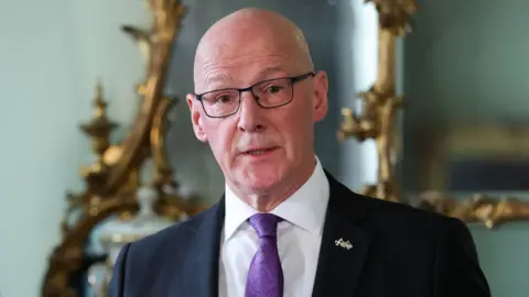 Getty Images John Swinney, who is bald with glasses, wearing a dark suit, white shirt and purple tie. He is speaking, with an ornamental mirror hanging on a wall behind him. 