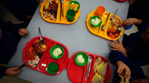 Four plastic trays, two red and two yellow, are seen from above in a school canteen. The trays are all full of food, including bowls of ice cream, sandwiches, and Sunday roasts. Children's hands can be seen reaching for the food.