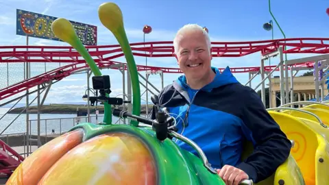 Derek Brockway on a carriage ride at Barry Island Pleasure Park, He has white hair and is wearing a blue and navy jacket