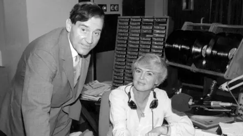 A black and white photo of Gloria with Alasdair in a radio news booth