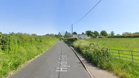 A Google street view screenshot of a country lane  with a hedge on one side and a field on the other. It's a sunny day and the sky is blue.