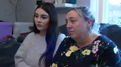 A young woman, possibly in her late teens, with long black hair, and a middle-aged woman with blonde hair tied in a bun, sit side by side on a sofa covered with a black plastic trash bag. The couple, mother and daughter, look worried as they look off camera.