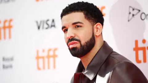 Drake poses on a red carpet at the Toronto International Film Festival. He is wearing a brown leather jacket.