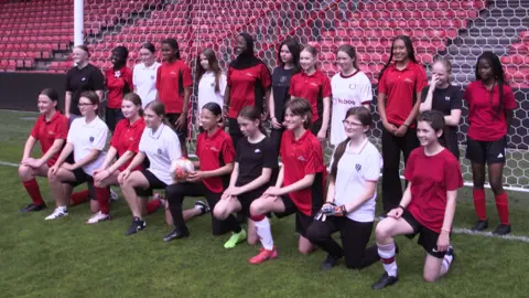 A group girls arranged in two lines in front of a goal, the front line kneeling and the second standing behind them. They are all smiling at a different camera to the left of the photographer, and there are rows of red stadium seating behind them. 