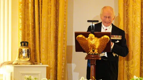 PA Media King Charles III wears a dark suit with a row of medals and a white shirt as he stands smiling behind a podium, behind him are golden curtains and next to him on a separate podium is a large golden bell engraved with the words TRUMP 1944. 