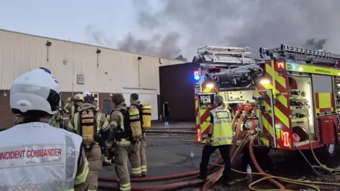 Northamptonshire Fire & Rescue The back views of seven firefighters looking towards a large white warehouse. Rising from it is black smoke. On the left, the firefighter is wearing a white helmet and tabard saying incident commander. On the right is the back of a fire engine with a firefighter in a yellow high vis tabard saying fire. 