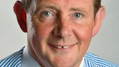 A headshot of Professor George Holmes, who is  smiling and wearing a white and blue stripped shirt.
