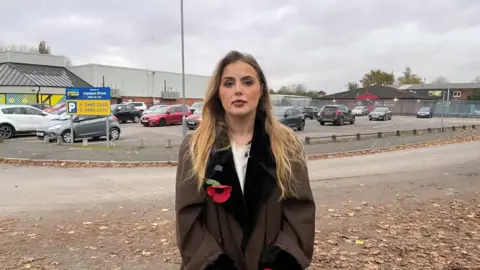 BBC Miss Hudson, who is a young blonde woman, stands outside the Excel carpark in question, wearing a brown coat with a poppy on the lapel