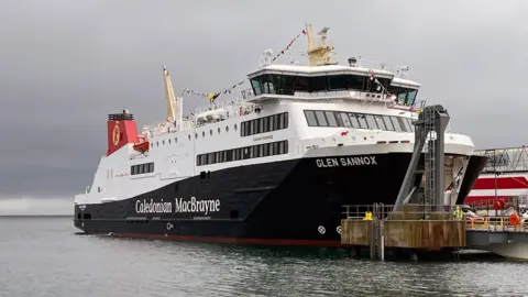 BBC Glen Sannox di samping dermaga di bawah langit fajar abu -abu. Perahu memiliki garis bendera di sepanjang bagian atas.