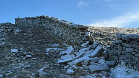 A granite stone wall rises up the side of a steep mountain. A section of the wall has collapsed and a large number of stones are lying in rubble.