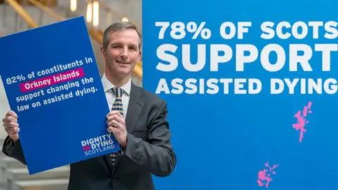 PA Media Liberal Democrat MSP Liam McArthur wears a suit and stands in front a sign which says "78% of Scots support assisted dying" and he holds a sign which says "82% of constituents in Orkney Islands support changing the law on assisted dying"