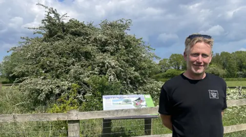 Edd Smith/BBC A man with a black T-shirt and blond hair smiles at the camera. He is standing in front of a tree which is behind a fence. An information panel is just behind the man but is not readable.