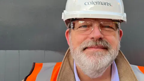 Ollie Conopo/BBC Man in a brown jacket with an orange hi-vis on top. He's wearing protective glasses and a hard hat.