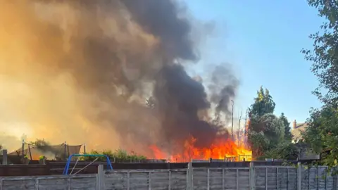 A picture of a garden fire taken a few rows back behind garden fencing. Orange flames have created a plume of dark smoke stretching upwards into the sky.