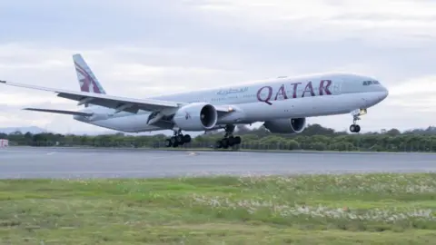 A wide shot of a plane taking off on a runway. "Qatar" is printed on the body of the plane.