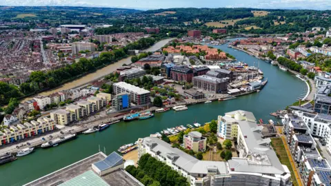 An aerial image capturing Bristol’s Harbourside area with the River Avon winding through the city. There are colourful residential houses and modern waterfront apartments, alongside a variety of boats docked along the quays. In the distance there are fields, hills and woodland. It looks like a warm and sunny day and the water is a deep green.