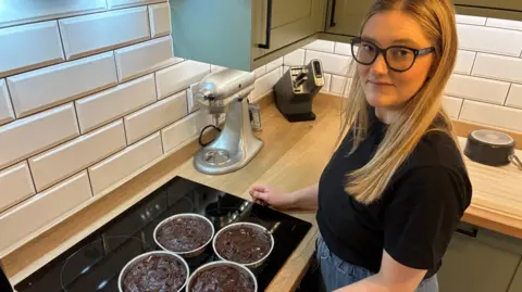 Laura Winter stands in the kitchen with the beginnings of chocolate brownie wreaths on a worktop in front of her.