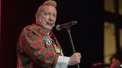 Dod Morrison John Lydon has short puni spiky ginger hair , a red and green tartan suit and holds a microphone stand.
