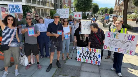 Protesters stand outside Hove Town Hall holding brightly-coloured signs opposing plans to close Wellington House day centre. Some of the signs say "every person matters" and "save our day centre". It's a sunny day with a blue sky and the protesters are wearing summer clothes and some have sunglasses.