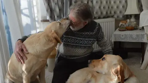 Contributed A man kneels on the floor in the middle of two golden Labrador dogs. One is licking his face as he smiles. The other is laid on the floor looking at them both. 
