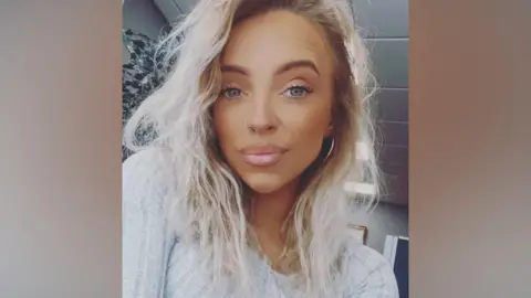 Natasha Gove, a woman with wavy, shoulder-length blonde hair, looks directly at the camera.  She is wearing a light grey ribbed jumper. 
