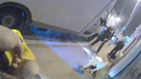 A grainy still from bodycam footage showing a man wearing shorts on the ground, two police officers beyond him and a hand carrying a Taser.