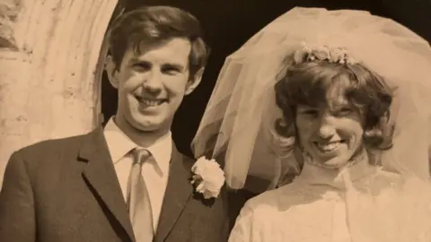 Slater family A black and white photo of a couple on their wedding day. The man is wearing a dark suit, white shirt and tie. The woman, in a white dress, has a large veil pushed back above her head. They are both smiling broadly, standing outside. 