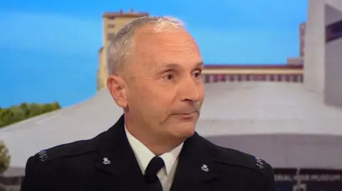 Mark Sigley, wearing police dress uniform, is sitting on the BBC Breakfast red sofa. Behind him is a studio backdrop that features images of Salford Quays in Manchester.