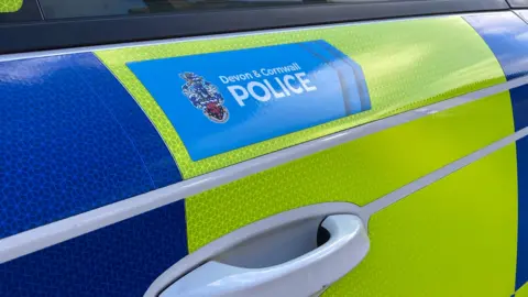 The door of a police car which is fluorescent yellow and blue and has the Devon and Cornwall Police logo which is as shield above the handle.