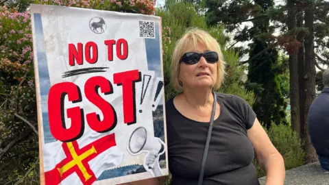 The image shows a woman holding a large "No to GST" placard. It bears a Guernsey flag. The woman is wearing black sunglasses and a black t-shirt with a strappy handbag over her shoulder.