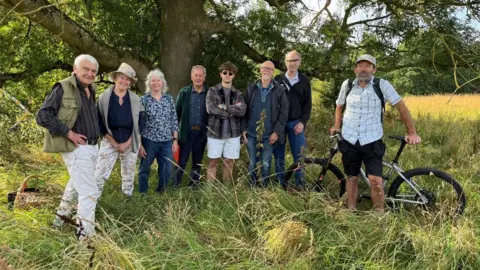 BBC/JULIA LEWIS Eight people stand in a field under a tree