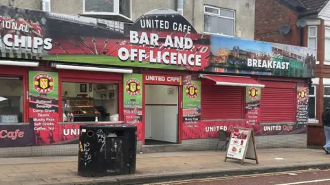 The outside of a café with a colourful fascia featuring images of Old Trafford stadium. The boards advertise food and drink and read 'United Cafe Bar and Off Licence'. 
