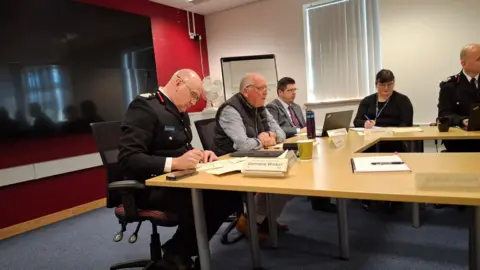 LDRS Men and women sit a desk during a meeting of the Shropshire & Wrekin Fire Authority. A TV can be seen in the background.