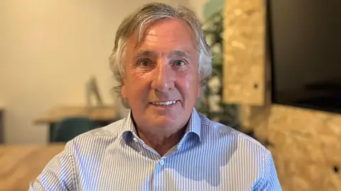 BBC A man with long grey hair in a striped shirt is sat on a chair with the rest of the room blurred in the background. 