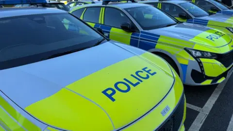 A close-up of a police car bonnet in line of marked police vehicles, parked in a row on a car park. 