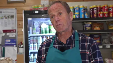 Peter Osborne is in a chequered shirt and blue apron. He is standing in front of food shelves and a fridge.