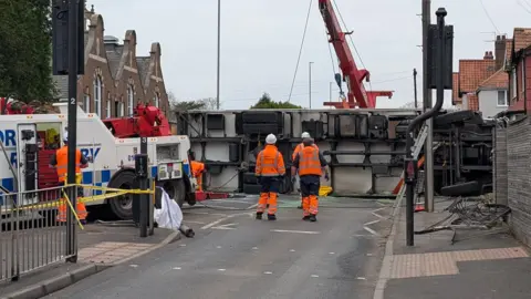 Jack Maclean/BBC An overturned lorry on its side on a residential street. A recovery team are pictured surrounding the vehicle.