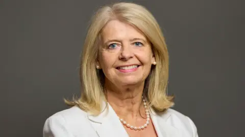 UK Parliament A woman with shoulder length blonde hair, a white jacked and pearl necklace