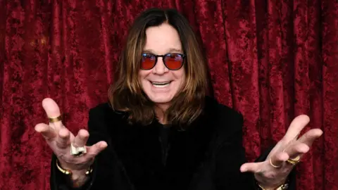 Getty Images A man with long brown hair and red sunglasses with his hands outstretched and a black top in front of a red curtain