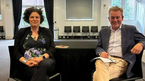 Claire Hanna and Mark Carruthers sit on chairs in a BBC production room and smile at the camera. She is wearing a black floral blouse and black suit and Mark wears cream chinos, a white shirt and a navy jacket and holds his glasses in his hand.