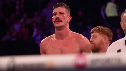 A sweaty topless man with short black hair and a moustache, stands in a boxing ring, with a rope visible in the foreground. A man is stood behind him looking to his right while a referee dressed in white stands next to him. 