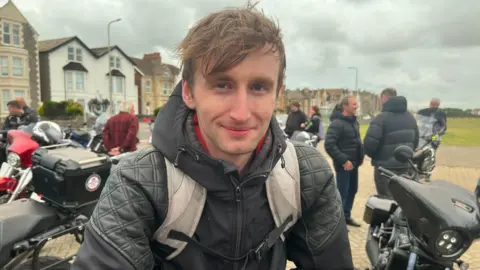 Mykel wearing a dark hoodie and a grey backpack with short light brown hair smiling at the camera. There are people loitering in the background in a car park with a few other motorbikes in view.