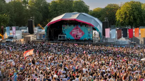 Ollie Ley A large festival crowd gathered looking at an empty stage which has "Bristol Pride" written on it. On either side there are speakers and colourful flags. Behind that are trees.