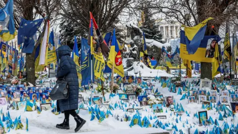 A woman dressed in an ankle-length winter coat walks at the snow-covered makeshift memorial to fallen Ukrainian defenders at Independence Square in Kyiv on a frosty winter day.