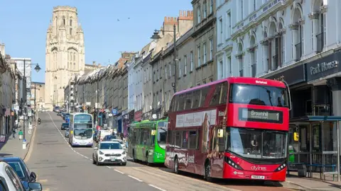 Getty Images The view up Park Street in Bristol towards the Wills Memorial Building, which is a tall tower in a gothic architectural style. There are rows of flats and shops up the hill, with buses and cars driving down it. 